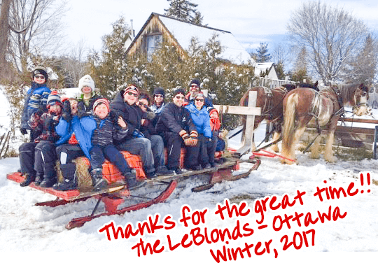 The LeBlond family with friends, enjoying a horse-drawn sleigh ride at Fallowfield Tree Farm - with a caption in red handwriting font that reads "Thanks for a great time!!, the Leblonds - Ottawa, Winter 2017"